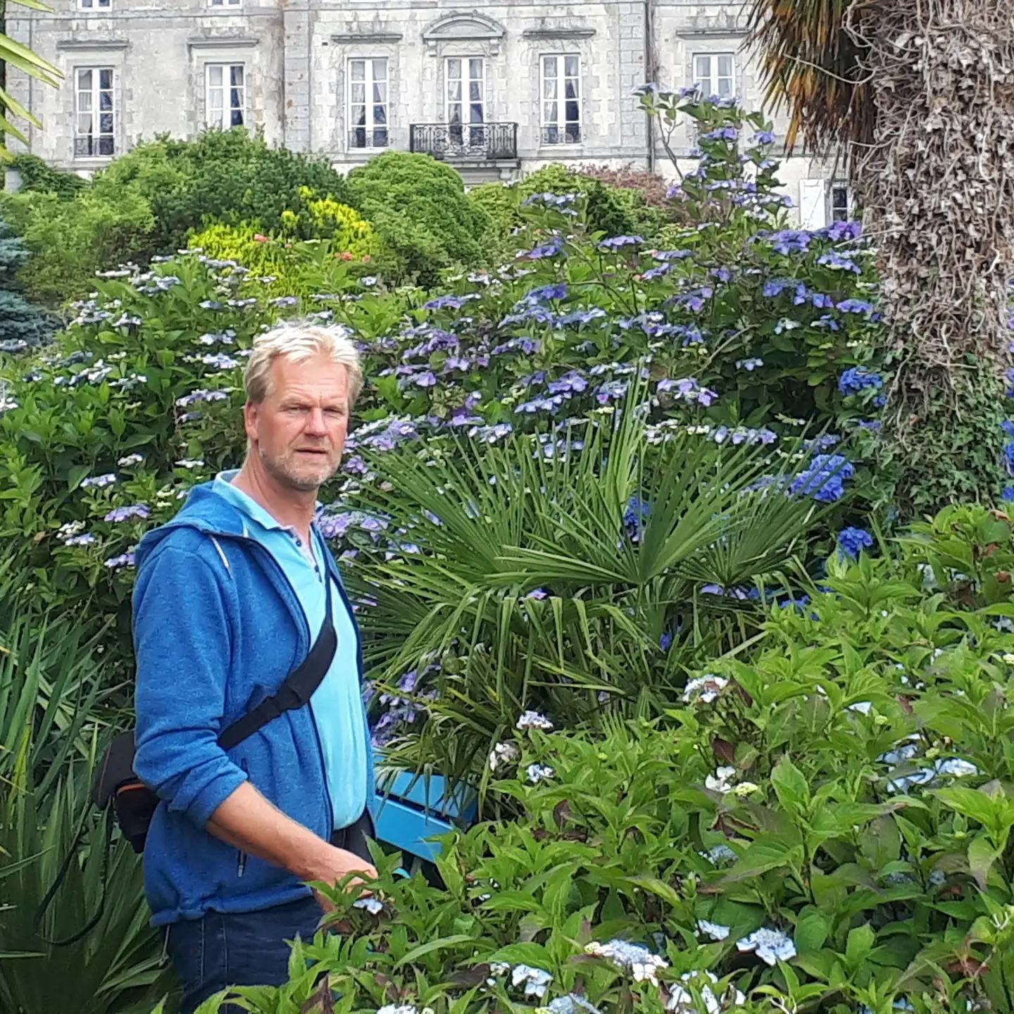 Marc Wilde in einer wundervoll angelegten grünen Gartenanlage mit Sträuchern,
 Pflanzen,
 Bäumen,
 im Hintergrund ein Herrenhaus